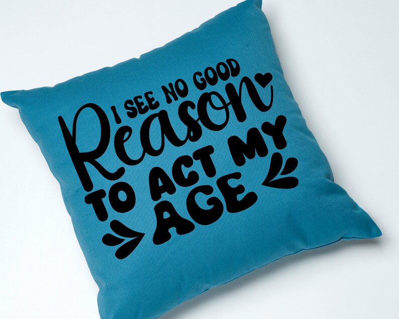 May include: A teal blue throw pillow with the text "I see no good reason to act my age" in black lettering with a small heart and leaf design.