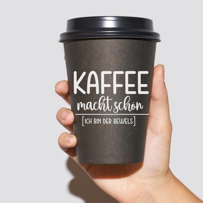 May include: A black paper coffee cup with a black lid. The cup has white text that reads "KAFFEE macht sch&ouml;n [ICH BIN DER BEWEIS]" which translates to "Coffee makes you beautiful [I AM THE PROOF]" in English.