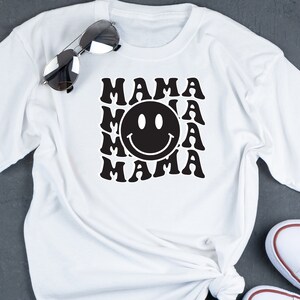 May include: White t-shirt featuring a black graphic design of the word "MAMA" repeated in a retro style with a smiley face in the centre.