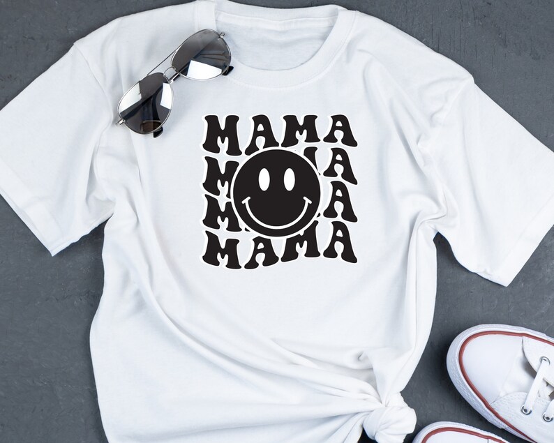 May include: White t-shirt featuring a black graphic design of the word "MAMA" repeated in a retro style with a smiley face in the centre.