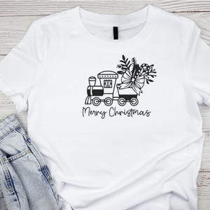 May include: White t-shirt with a black and white graphic of a toy train with a Christmas tree and bow on top. The text "Merry Christmas" is below the train.