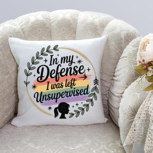 May include: A white square pillow with the text "In my defense I was left unsupervised" in a circular design. The design includes a rainbow stripe, stars, and a silhouette of a person. Green leaves and branches surround the text.