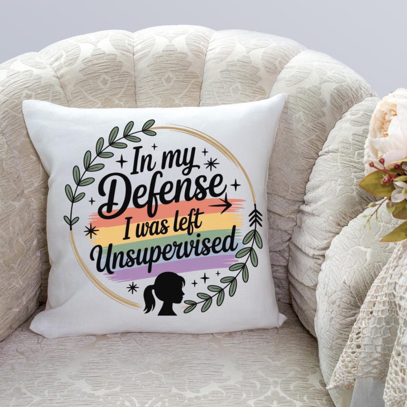 May include: A white square pillow with the text "In my defense I was left unsupervised" in a circular design. The design includes a rainbow stripe, stars, and a silhouette of a person. Green leaves and branches surround the text.