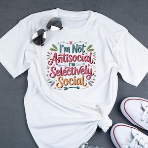 May include: White t-shirt with the text "I'm Not Antisocial, I'm Selectively Social" in colorful, decorative lettering. Sunglasses rest on the shirt. White sneakers are visible in the bottom right corner. A casual, everyday wear tee.
