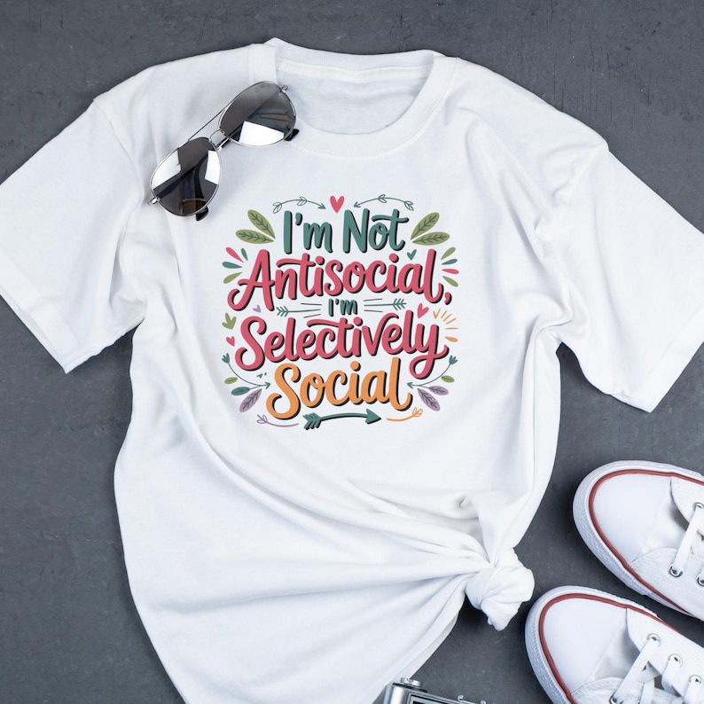 May include: White t-shirt with the text "I'm Not Antisocial, I'm Selectively Social" in colorful, decorative lettering. Sunglasses rest on the shirt. White sneakers are visible in the bottom right corner. A casual, everyday wear tee.