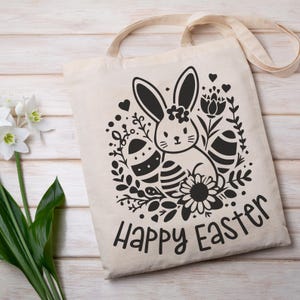 May include: A white tote bag featuring a black and white illustration of a bunny rabbit holding an Easter egg. The bunny is surrounded by flowers and the text "Happy Easter" is printed below the illustration.