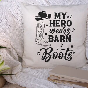 以下が含まれることがあります： 黒い文字で「My hero wears barn boots」と書かれた白いクッション。ブーツの言葉の前後に音符が記されている。クッションには、黒いカウボーイハットと黒いカウボーイブーツも描かれている。