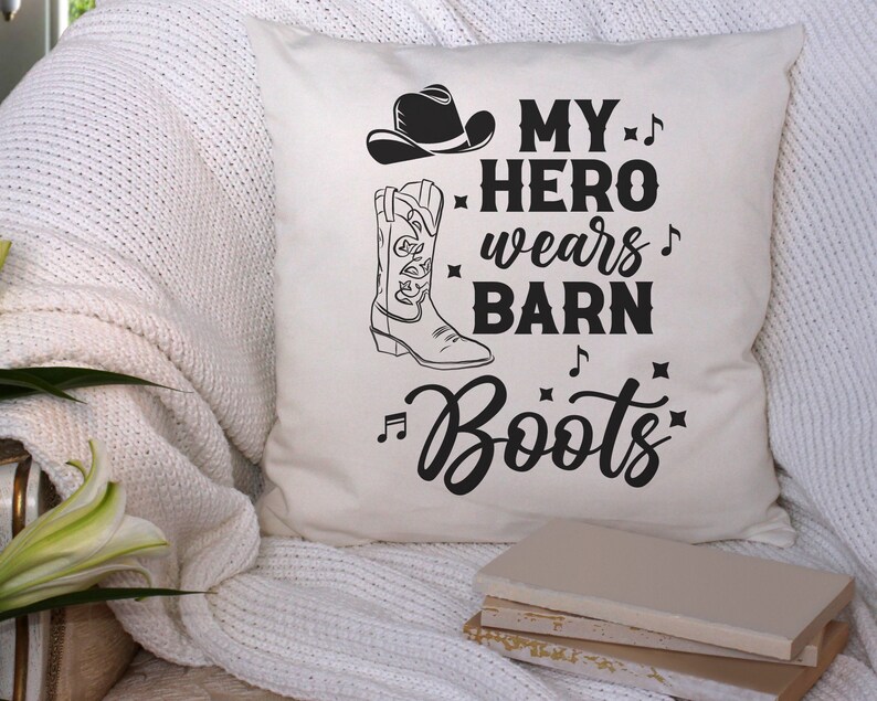 以下が含まれることがあります： 黒い文字で「My hero wears barn boots」と書かれた白いクッション。ブーツの言葉の前後に音符が記されている。クッションには、黒いカウボーイハットと黒いカウボーイブーツも描かれている。