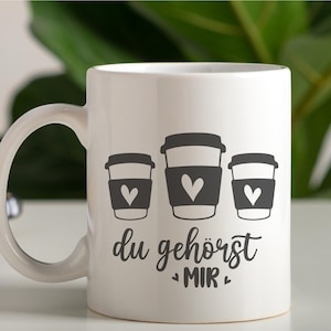 May include: White ceramic coffee mug featuring three stylized coffee cup graphics with heart-shaped designs. The mug has the German text "du geh&ouml;rst MIR" in a script font.