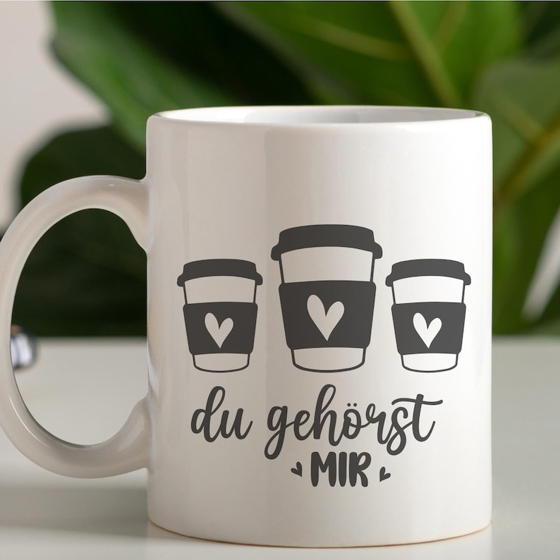 May include: White ceramic coffee mug featuring three stylized coffee cup graphics with heart-shaped designs. The mug has the German text "du geh&ouml;rst MIR" in a script font.