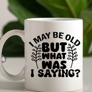 May include: White ceramic mug with a black design that reads "I may be old but what was I saying?" The design includes two stylized plant sprigs.
