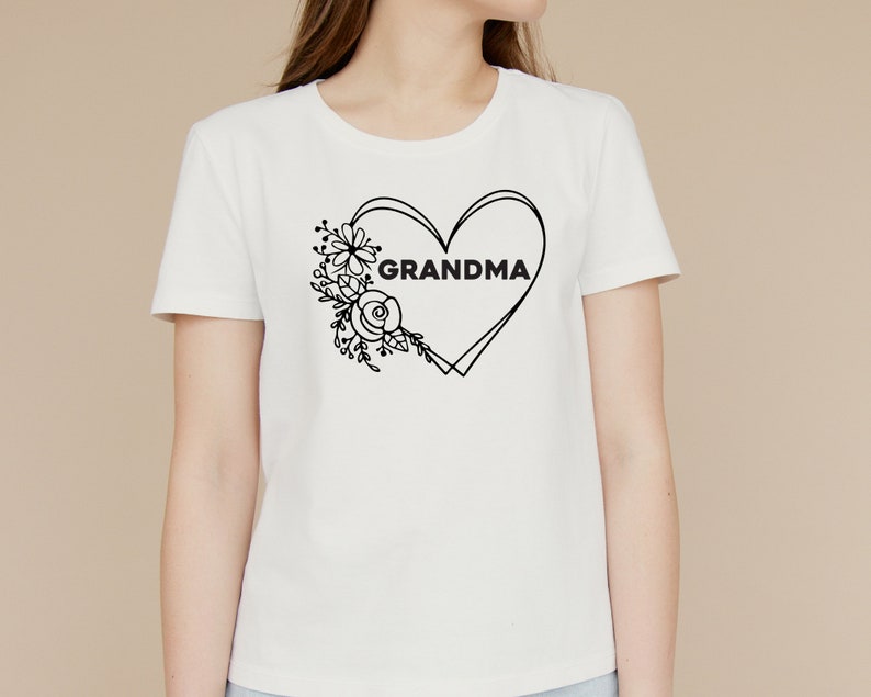 May include: White t-shirt with a black outline of a heart with the word "GRANDMA" inside. The heart is surrounded by a floral design.