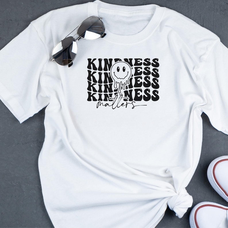 May include: White t-shirt with a black graphic design that says "Kindness matters" in a retro font. A black smiley face with dripping eyes is centered above the text.