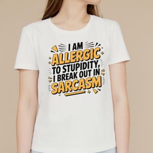 May include: White t-shirt with the text "I AM ALLERGIC TO STUPIDITY, I BREAK OUT IN SARCASM" in black and yellow. The shirt has a crew neck and short sleeves. The text is in a bold, stylized font.