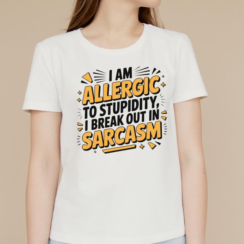 May include: White t-shirt with the text "I AM ALLERGIC TO STUPIDITY, I BREAK OUT IN SARCASM" in black and yellow. The shirt has a crew neck and short sleeves. The text is in a bold, stylized font.