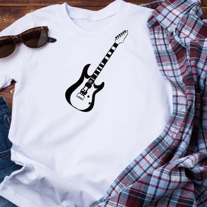 May include: White t-shirt featuring a black electric guitar graphic. The guitar design is a simple outline. Sunglasses and a plaid shirt are also visible in the image. The shirt is made of a soft material.