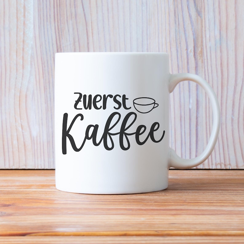 May include: White ceramic coffee mug with the words "Zuerst Kaffee" in black script. A small coffee cup graphic is above the word "Kaffee". The mug has a curved handle and is set against a light wood background.