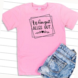 Op de afbeelding: Roze T-shirt met zwarte tekst die "Wein gut. Alles GUT." luidt. De tekst is ingelijst in een zwart vierkant met een klein hartje en bladeren onderaan.