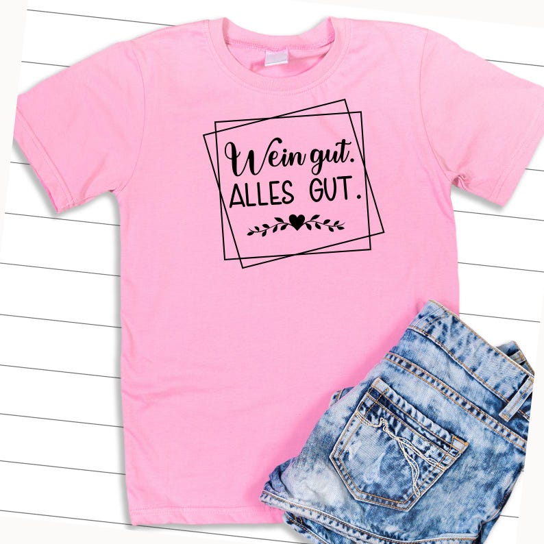 Op de afbeelding: Roze T-shirt met zwarte tekst die "Wein gut. Alles GUT." luidt. De tekst is ingelijst in een zwart vierkant met een klein hartje en bladeren onderaan.