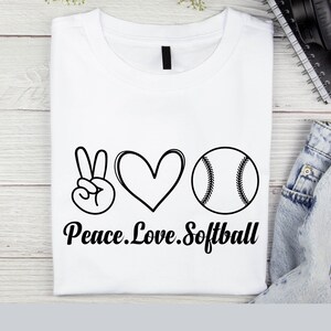 May include: White t-shirt with a peace sign, heart, and softball graphic. The text "Peace. Love. Softball" is printed below the graphic.