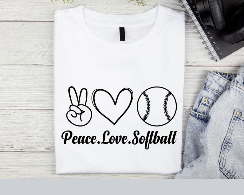 May include: White t-shirt with a peace sign, heart, and softball graphic. The text "Peace. Love. Softball" is printed below the graphic.