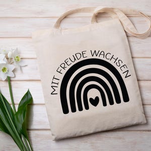 Peut inclure: Un sac fourre-tout en toile blanche avec du texte noir qui dit "Mit Freude Wachsen" et un arc-en-ciel noir avec un c&oelig;ur au centre.