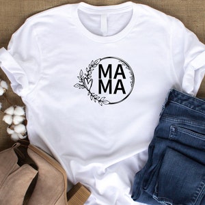 May include: White t-shirt with a black circle design featuring the word "MAMA" in all caps and a floral wreath around the circle.