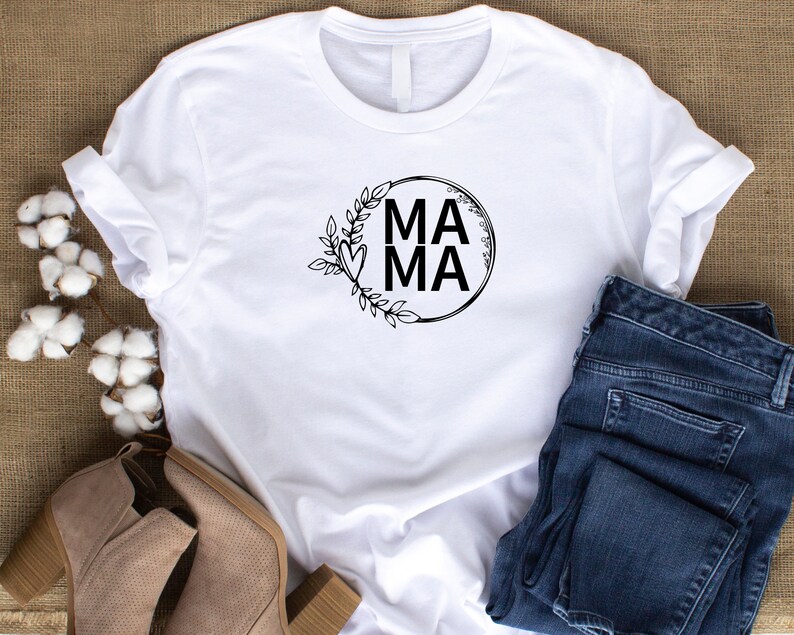 May include: White t-shirt with a black circle design featuring the word "MAMA" in all caps and a floral wreath around the circle.