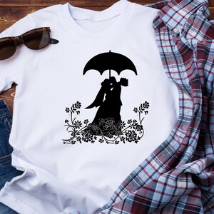 May include: White t-shirt with a black silhouette of a bride and groom under an umbrella. The couple is surrounded by floral designs.