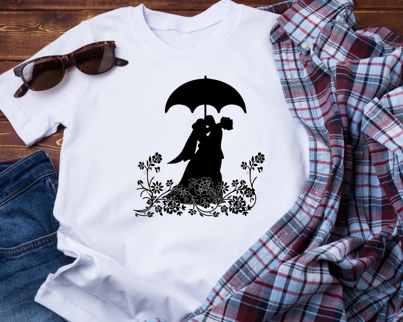 May include: White t-shirt with a black silhouette of a bride and groom under an umbrella. The couple is surrounded by floral designs.