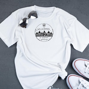 May include: White t-shirt with a black graphic design of a circle with a wreath and the text "Willkommen in unserem Zuhause" and a row of houses.