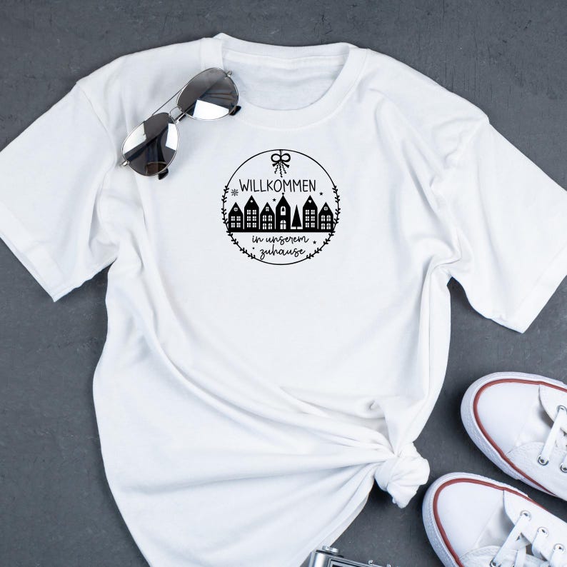 May include: White t-shirt with a black graphic design of a circle with a wreath and the text "Willkommen in unserem Zuhause" and a row of houses.