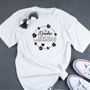 Peut inclure: T-shirt blanc avec un texte noir qui dit "Danke fur die tolle Kindergartenzeit" entour&eacute; d'un cercle de fleurs noires.