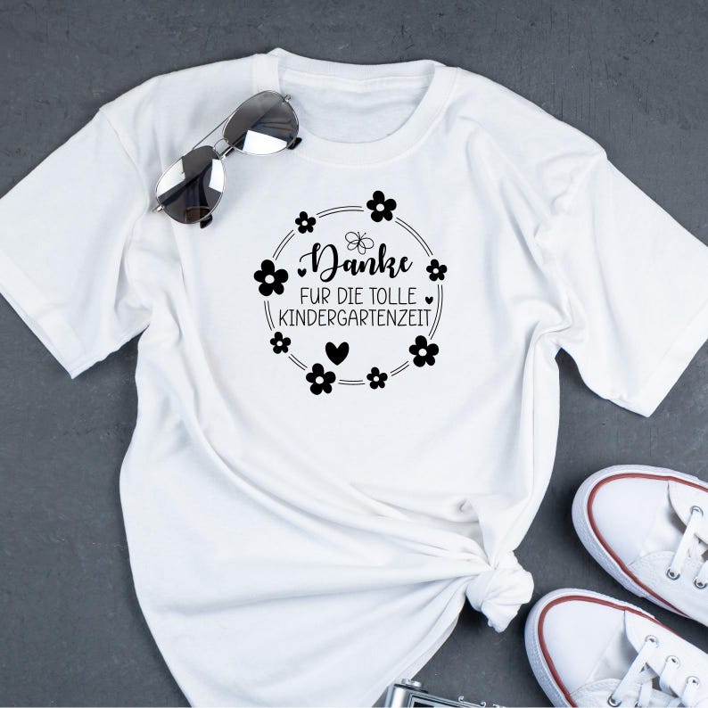 Peut inclure: T-shirt blanc avec un texte noir qui dit "Danke fur die tolle Kindergartenzeit" entour&eacute; d'un cercle de fleurs noires.