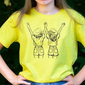 May include: A yellow t-shirt with a black graphic of two girls wearing cowboy hats and shorts, with their arms raised in the air.