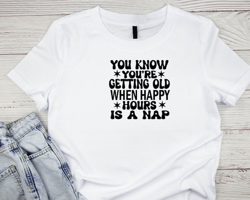 May include: White t-shirt with black text that reads "YOU KNOW *YOU'RE* GETTING OLD WHEN HAPPY *HOURS* IS A NAP"