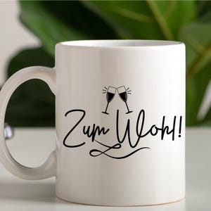 Op de afbeelding: Witte keramische mok met een zwart ontwerp van twee klinkende wijnglazen en de tekst "Zum Wohl!"