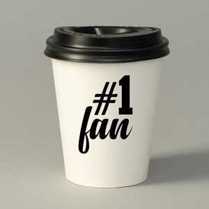 May include: White to-go coffee cup with a black lid. The cup has black text that says "#1 fan".