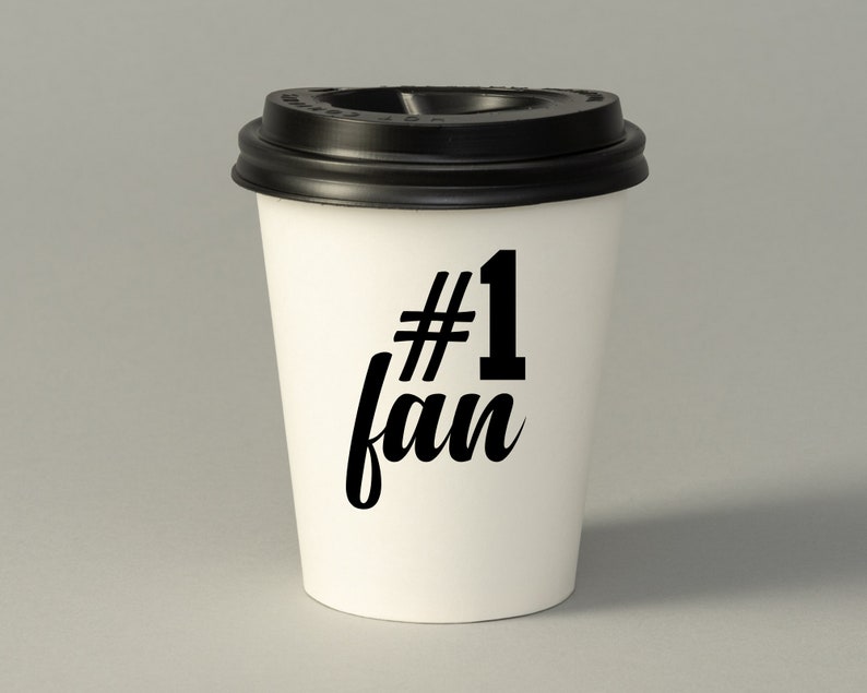 May include: White to-go coffee cup with a black lid. The cup has black text that says "#1 fan".