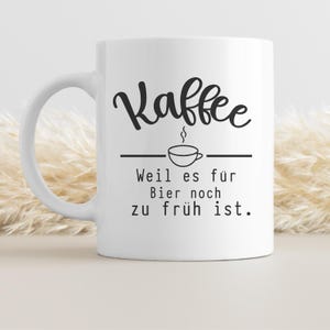 May include: White ceramic coffee mug with a black script font that reads "Kaffee" above a line drawing of a coffee cup. Below the cup, the text reads "Weil es f&uuml;r Bier noch zu fr&uuml;h ist."
