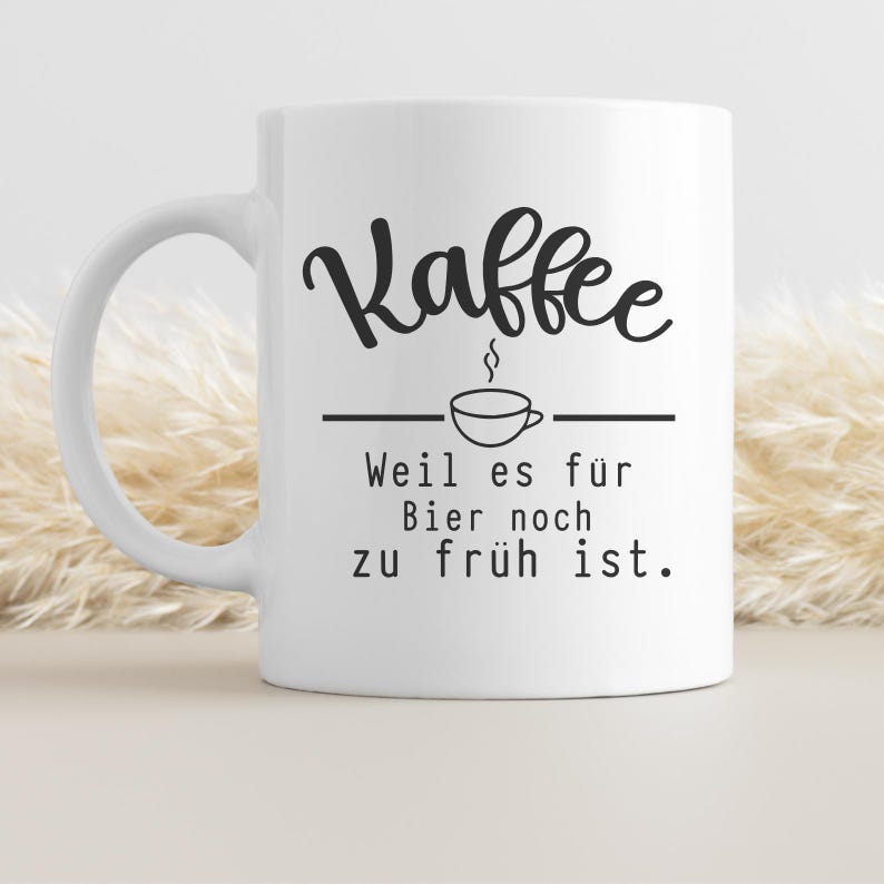 May include: White ceramic coffee mug with a black script font that reads "Kaffee" above a line drawing of a coffee cup. Below the cup, the text reads "Weil es f&uuml;r Bier noch zu fr&uuml;h ist."