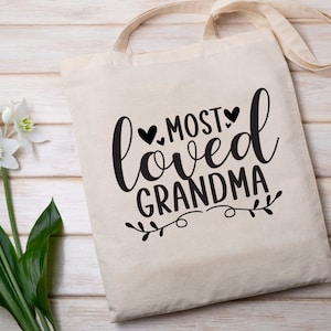 May include: A white canvas tote bag with black text that reads "Most Loved Grandma" with a small floral design and three black hearts.