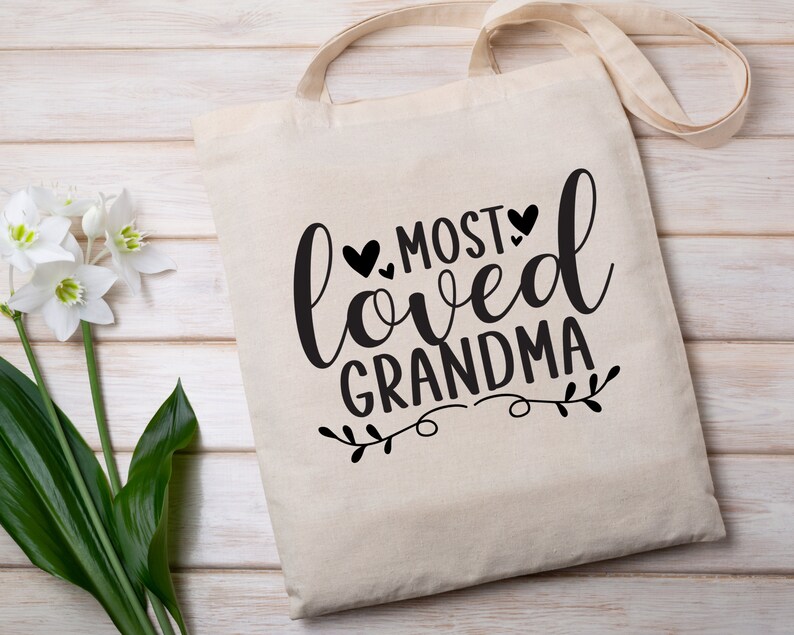 May include: A white canvas tote bag with black text that reads "Most Loved Grandma" with a small floral design and three black hearts.
