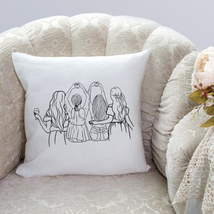 May include: A white throw pillow with a black line drawing of four women standing together. The women are facing away from the viewer and have their arms around each other. Two of the women are making heart shapes with their hands.