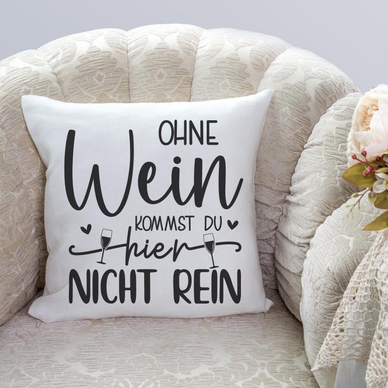 Op de afbeelding: Een wit kussen met zwarte tekst die "Ohne Wein Kommst Du Hier Nicht Rein" luidt, wat vertaald wordt als "Zonder wijn kom je hier niet binnen".