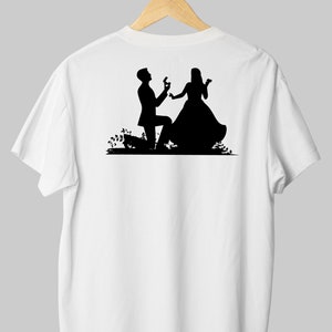 May include: A white t-shirt with a black silhouette of a couple. The man is on one knee, holding a ring, and the woman is smiling. The silhouette is set against a simple background of flowers.