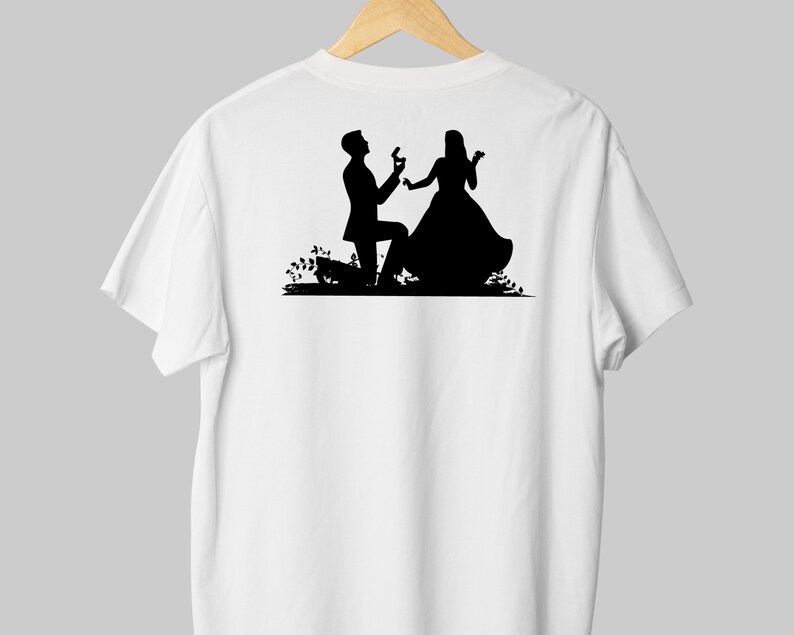 May include: A white t-shirt with a black silhouette of a couple. The man is on one knee, holding a ring, and the woman is smiling. The silhouette is set against a simple background of flowers.