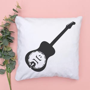 May include: White square pillow with a black guitar silhouette design. The guitar has a unique circular design on the body. The pillow is on a pink surface with green leafy branches in the corner.