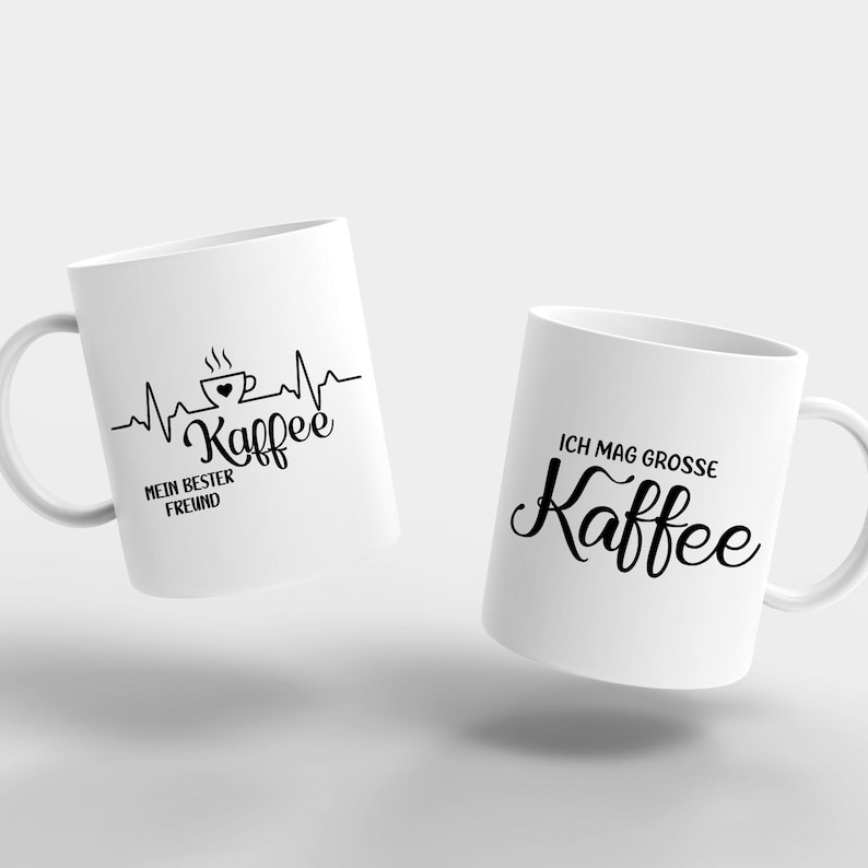 May include: Two white ceramic coffee mugs. One mug features a black graphic of a coffee cup with a heartbeat line and the text "Kaffee" and "MEIN BESTER FREUND". The other mug has the text "ICH MAG GROSSE Kaffee".