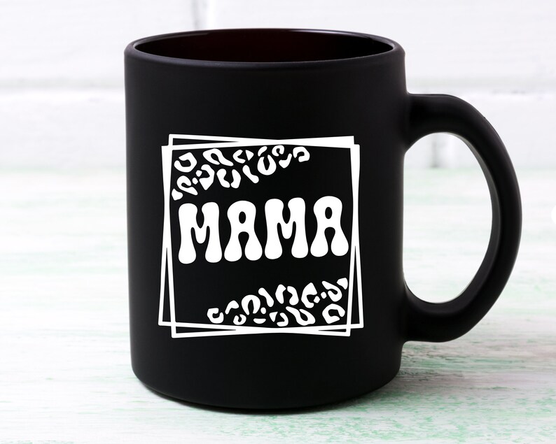 May include: Black ceramic coffee mug with a white design of a leopard print frame around the word "MAMA" in a retro font.